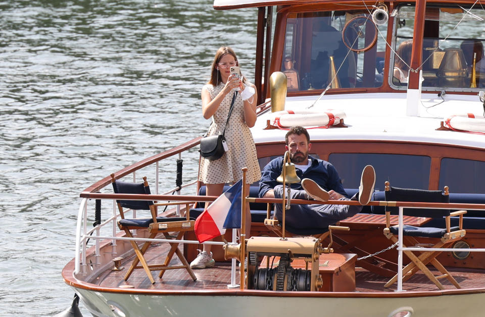 Jennifer Lawrence and Adam Sandler are on a boat. Jennifer is standing and taking a photo while Adam is lying with his feet up