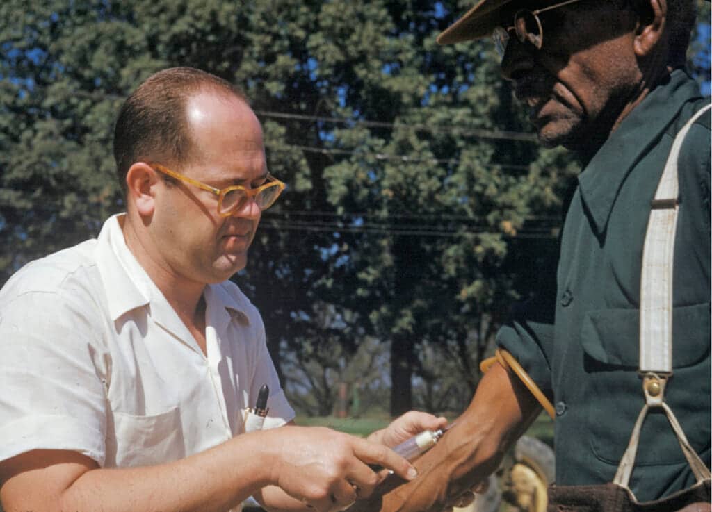 FILE – In this 1950’s photo released by the National Archives, a Black man included in a syphilis study has blood drawn by a doctor in Tuskegee, Ala. (National Archives via AP, File)