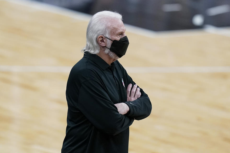 Gregg Popovich wears a face mask and has his arms crossed on the sideline of a game.