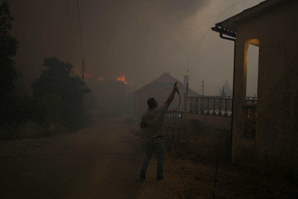 An man uses a garden hose to water the roof of a house as a forest fire smoke darkens the sky in the village of Bemposta, near Ansiao, central Portugal, Wednesday, July 13, 2022. Thousands of firefighters in Portugal continue to battle fires all over the country that forced the evacuation of dozens of people from their homes. (AP Photo/Armando Franca)