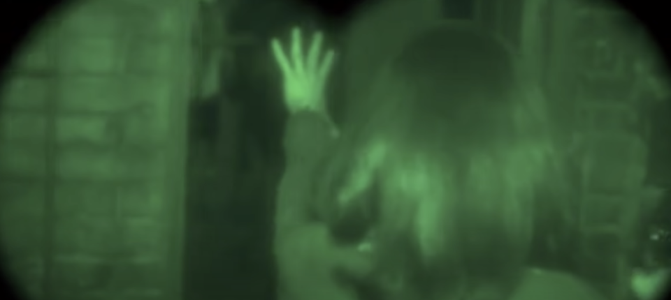 A woman walks blindly through a dark home, being followed by a man wearing night googles