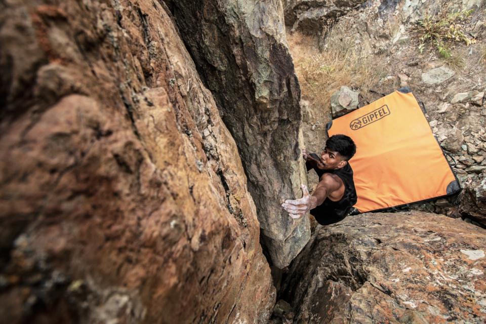 A top down view of a man climbing a boulder. Beneath him, an orange crash pad is visible to protect him from falling onto the rocky terrain below.