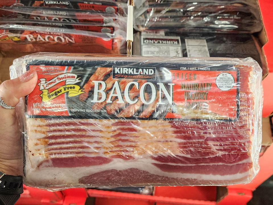 A hand holds a package of Kirkland Signature bacon with a red and black label in front of a display