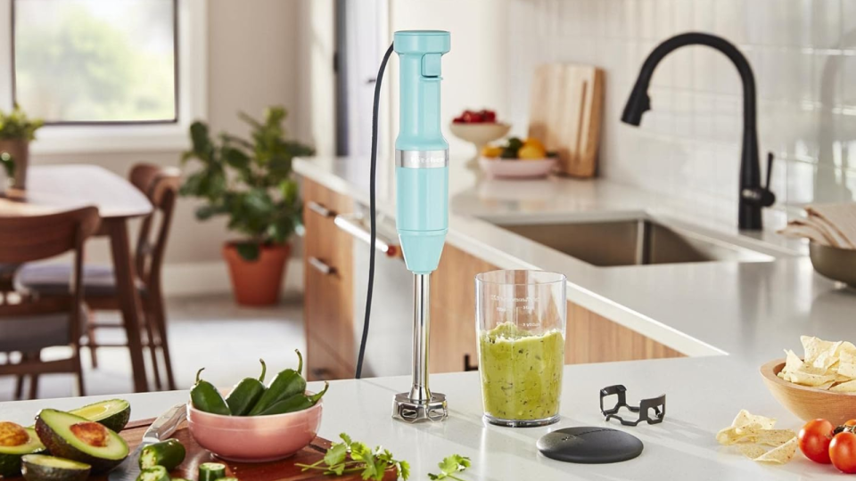 the teal kitchenaid immersion blender on a kitchen counter next to a container of guacamole and surrounded by various ingredients