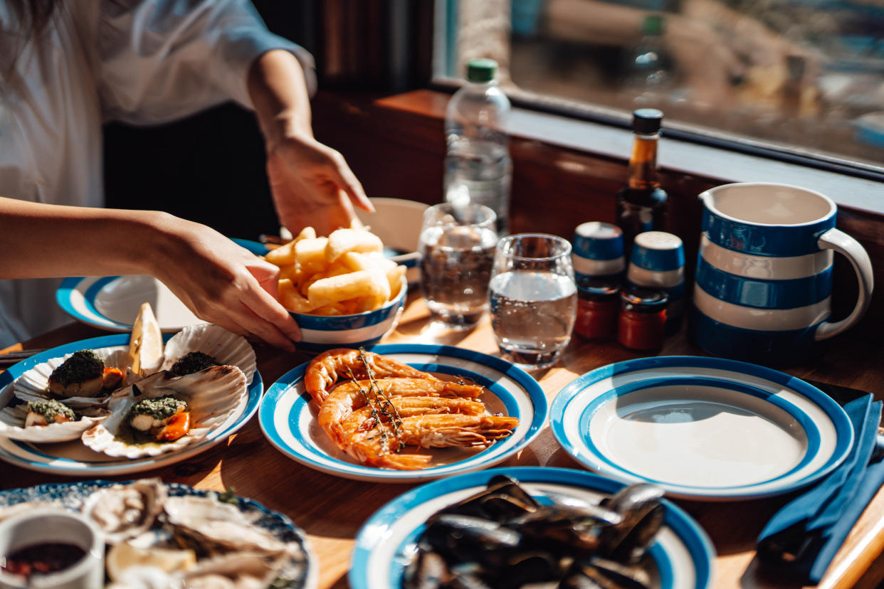 Cropped shot of female hands passing a bowl of French fries, various seafood served on the dining table with beautiful sunlight. Sharing and togetherness concept.