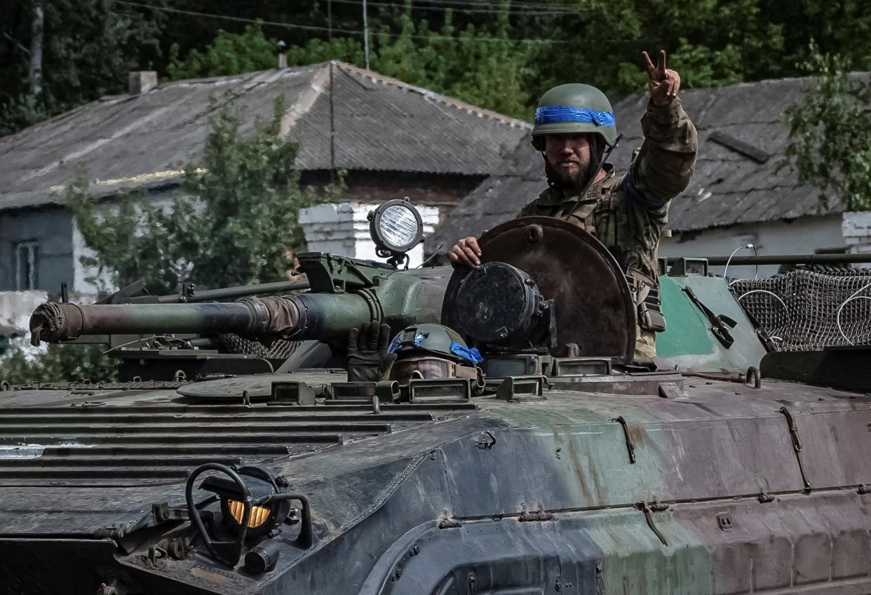 Two Ukrainians in an infantry fighting vehicle. One is holding a peace sign, houses in the background.