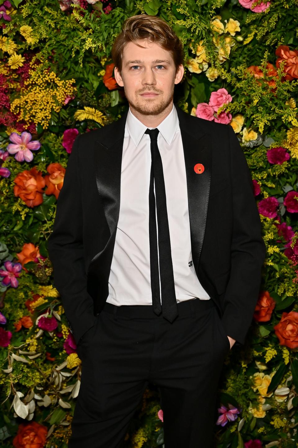 Joe Alwyn stands in front of a floral backdrop wearing a black suit and tie with a red poppy pin