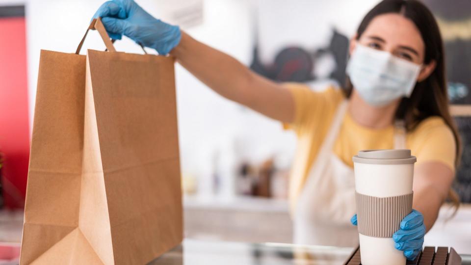 Young woman wearing face mask while serving takeaway breakfast and coffee inside cafeteria restaurant - Worker preparing delivery food inside bakery bar during coronavirus period - Focus on right hand.