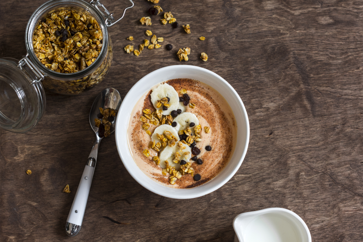 Top view of yogurt with granola and Nutella in a white bowl, next to a spoon, surrounded by a glass jar of granola and a white jug of milk on a dark wooden table