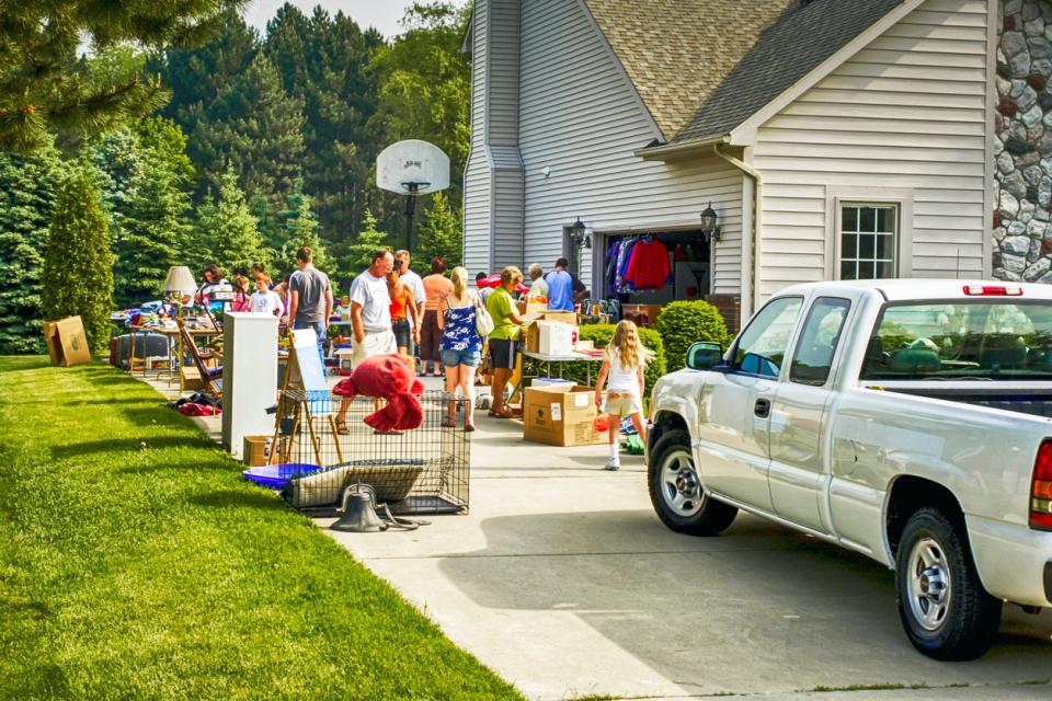 driveway with pickup truck parked and tables set up for yard sale and groups of people shopping outside large house