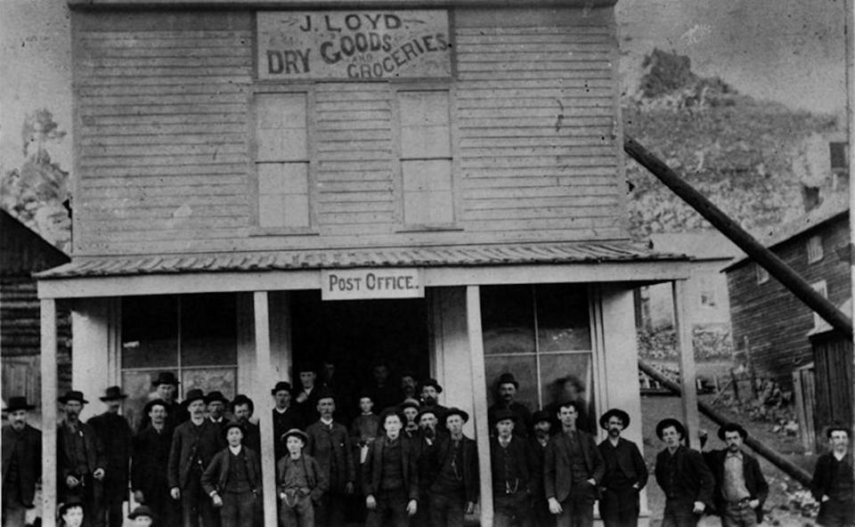 Men and boys standing on the porch of the J. Loyd Dry Goods and Groceries business. A Post Office sign hangs over the entrance.