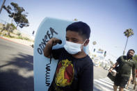 Elijah Cobos wear a face mask as he carries a boogie board to the beach Sunday, July 12, 2020, in Santa Monica, Calif., amid the coronavirus pandemic . A heat wave has brought crowds to California's beaches as the state grappled with a spike in coronavirus infections and hospitalizations. (AP Photo/Marcio Jose Sanchez)