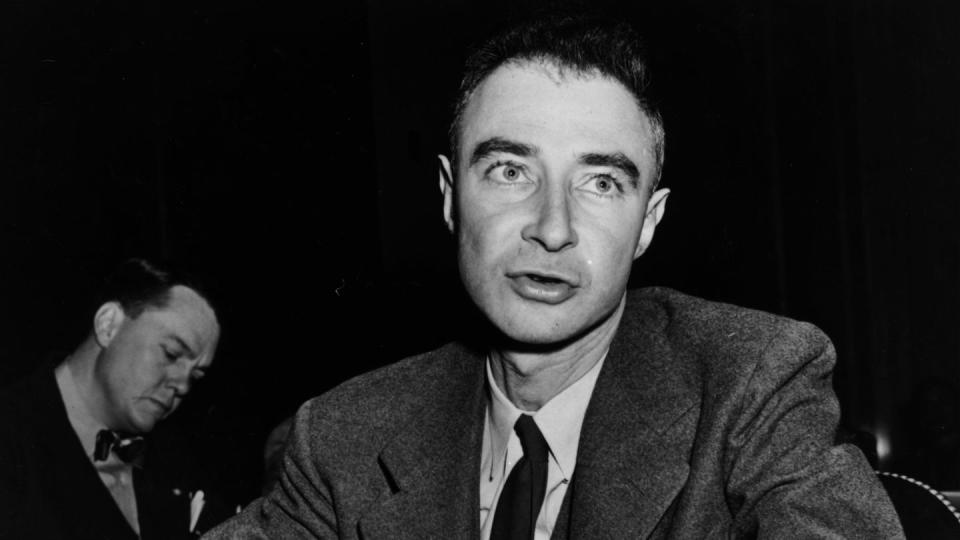 a black and white photo of j robert oppenheimer, wearing a suit and sitting at a table, speaking to someone off camera