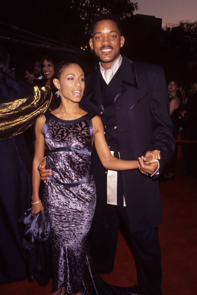 the couple on red carpet; Jada on left in patterned dress with updo hairstyle, Will on right in long coat holding her hand