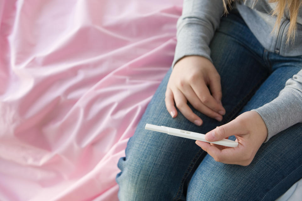Person sitting on a bed, holding a pregnancy test, wearing jeans and a gray long-sleeve shirt. Faces are not visible