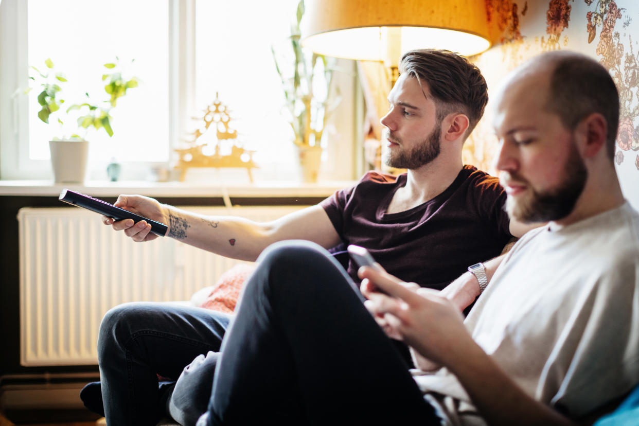 A gay couple relaxing at home together, watching tv and using a smartphone.