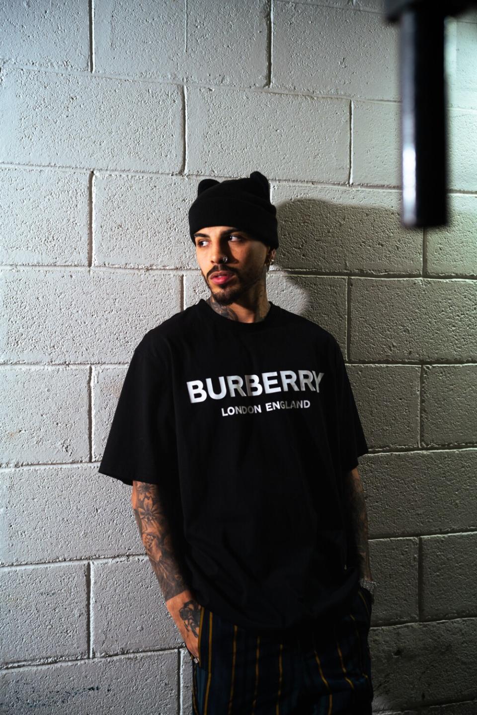 A man in a black skip cap and black T-shirt, in front of a white cinder block wall