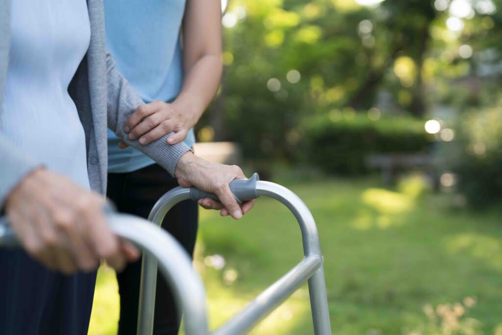 Image of an unrecognizable senior woman being assisted to walk using a walker by caregiver at park.