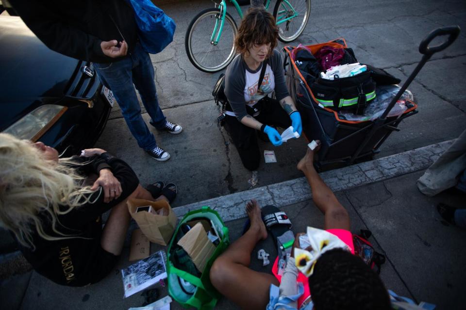 A woman wearing gloves gives first aid to a woman on the sidewalk with an open wound on her foot.