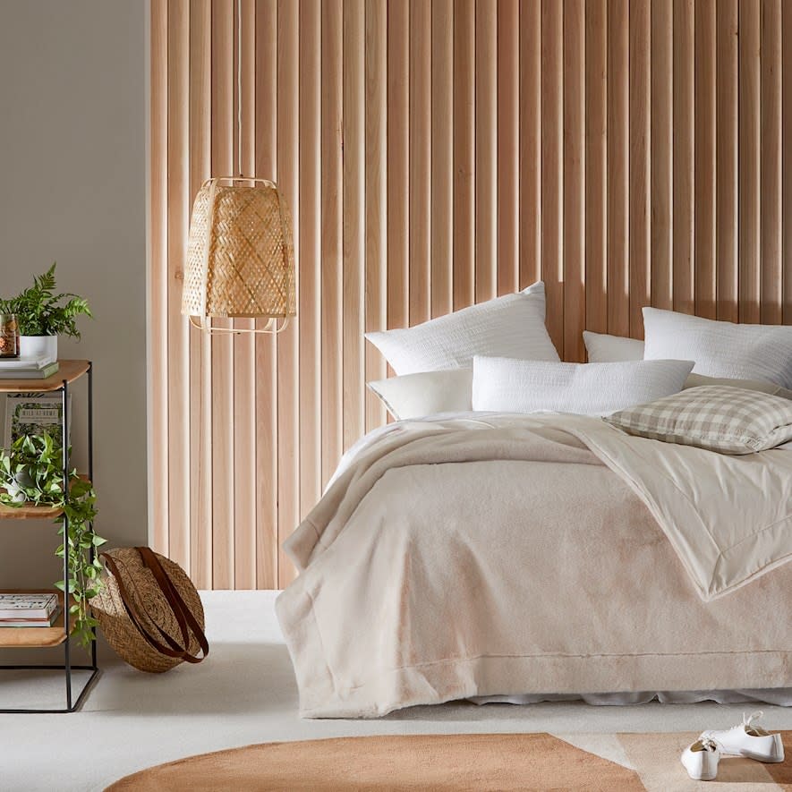 A neutral toned room featuring wicker hanging lantern, in door plants, wood toned blinds and bed with white pillows and soft beige fur blanket