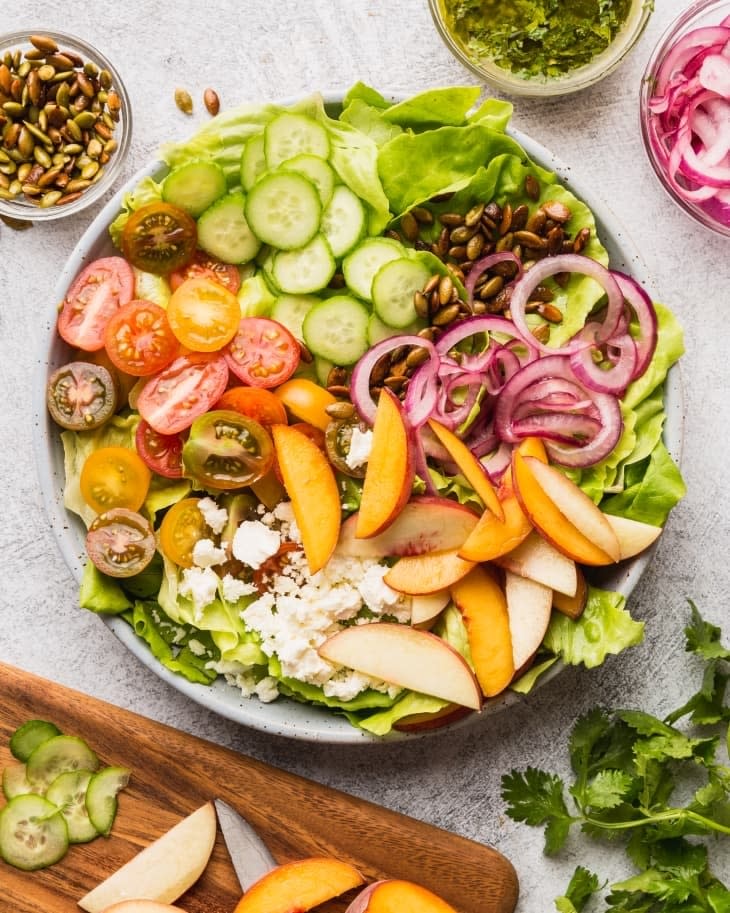 Plate of salad with cucumbers, cherry tomatoes, red onions, seeds, feta cheese, and peach slices; ingredients and dressing in bowls nearby