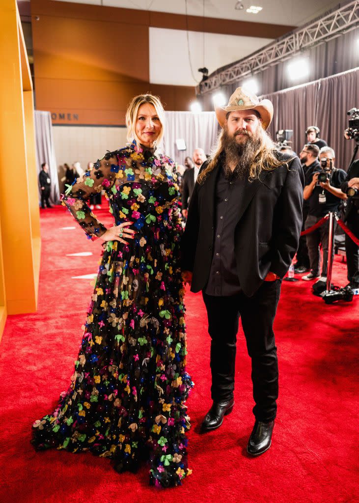 morgan stapleton and chris stapleton stand on a red carpet and post for a photo, she has one hand on her hip and wears a black dress with multicolored applique flowers, he wears a light brown cowboy hat with a black suit and shirt