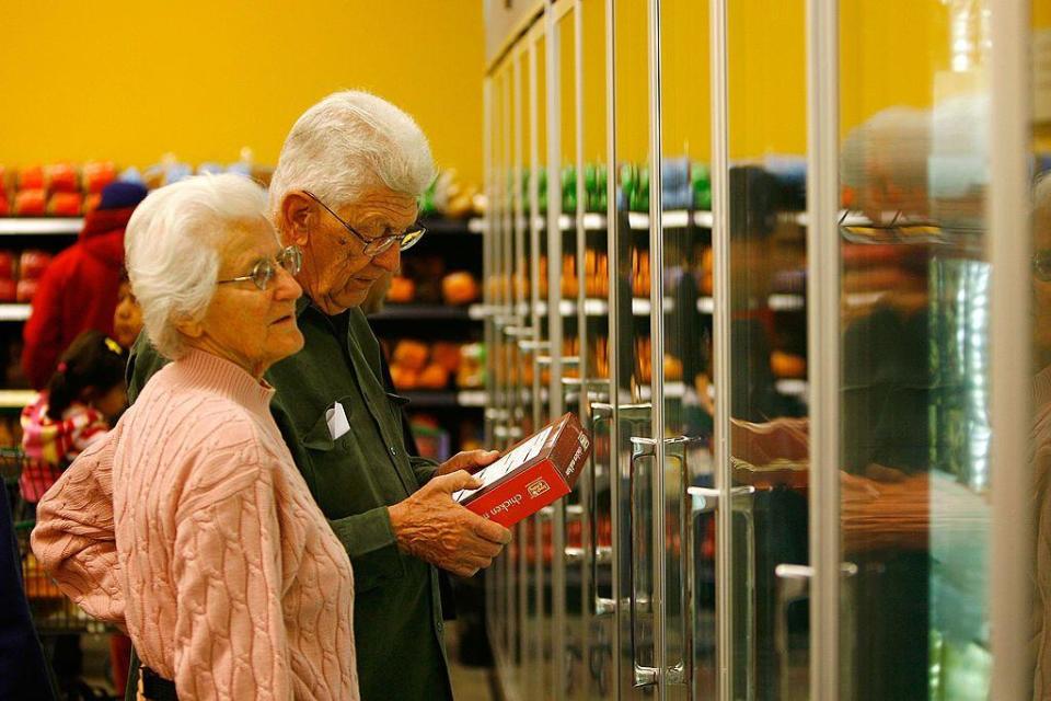Pareja de ancianos con una caja de alimentos ya preparados frente a los refrigeradores de un supermercado en Estados Unidos.