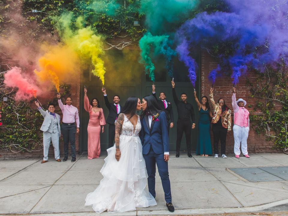 Two brides kiss as rainbow smoke bombs explode behind them.