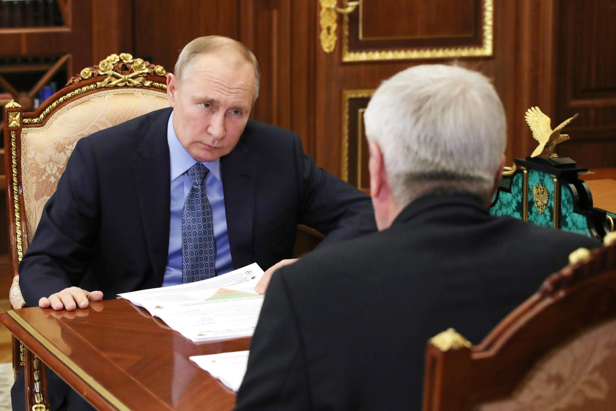 Russian President Vladimir Putin, seated in an ornate chair with hands on a wooden table, leans forward and scowls at head of Russia's Federal Service for Financial Monitoring Yuri Chikhanchin, seated across from him and seen from behind