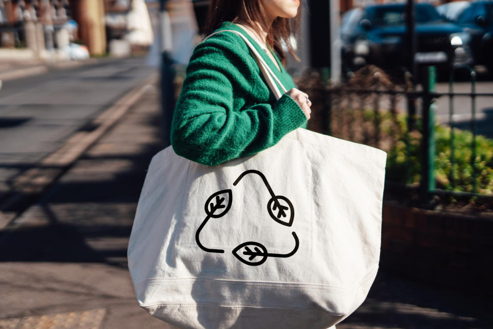 Woman carrying a large tote bag with a recycle symbol and leaves on it, walking on a sidewalk