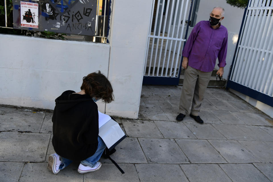 A student looks for his document stating a negative COVID-19 self-test as a teacher waits at the entrance of the school in Athens, Monday, May 10, 2021. More than 1.1 million pupils and 127,300 teachers returned to school on Monday as primary and junior high school reopened its doors with mandatory home self-tests. (AP Photo/Michael Varaklas)