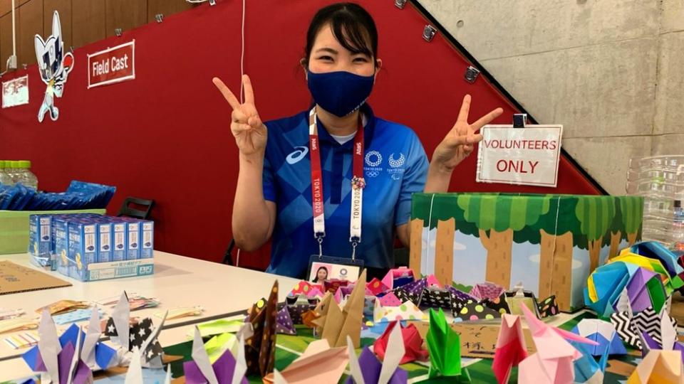 A volunteer doing the victory sign, sitting behind a table with origami.