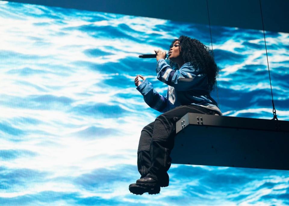 sza sits on the end of a hanging platform with her legs hanging off the end, she sings into a microphone she holds in one hand, behind her is a large screen projecting waves, she wears a long sleeve blue jersey style shirt, black cargo pants, and black boots