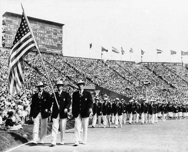 FILE - This is a July 29, 1948 file photo of Ralph Craig, center, of Albany, N.Y., who won the 100- and 200-meter dashes in the 1912 Olympic games in Stockholm, and is a member of the U.S. Olympic Yachting team in the 1948 games, carrying the American flag in the parade of the nations at the opening of the Olympic games in London's Wembley Stadium. Athletes bought their own uniforms, and some their own food. They stayed in private homes, schools and military barracks. If eggs appeared on the training menu, it was a cause for celebration. When London hosted the Olympics in 1948, organizers did it on the cheap, and they made no apologies about it. (AP Photo/File)