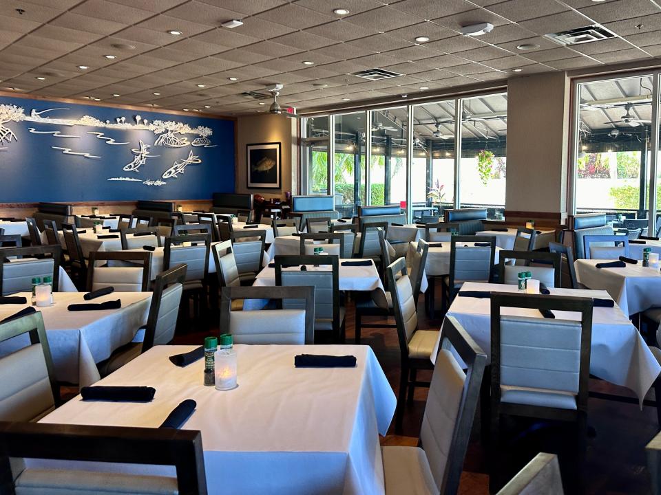 A dining area filled with square tables with white tablecloths and chairs with black frames and white cushions. A blue design in the back of the room depicts white illustrations of fish