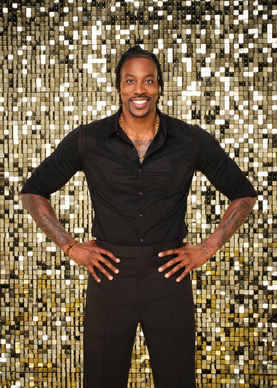 Dwight Howard poses with hands on hips in front of a glittery backdrop, wearing a stylish black outfit