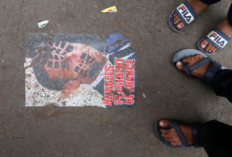 People stand next to a defaced poster of France's President Emmanuel Macron on a road as a mark of a protest, in Mumbai