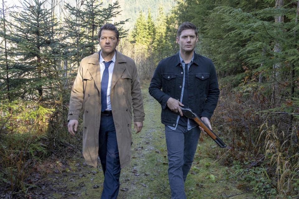 Misha Collins, wearing a trench coat and tie, and Jensen Ackles, in casual clothes with a shotgun, walk through a forest in a scene from "Supernatural"