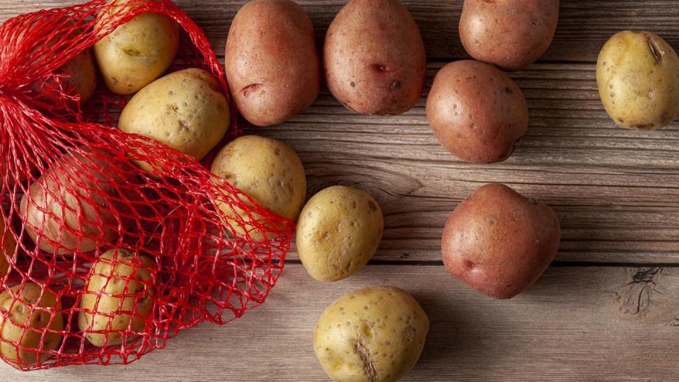 flat lay close up image featuring a red mesh potato sack with pink and yellow raw organic potatoes on wooden background