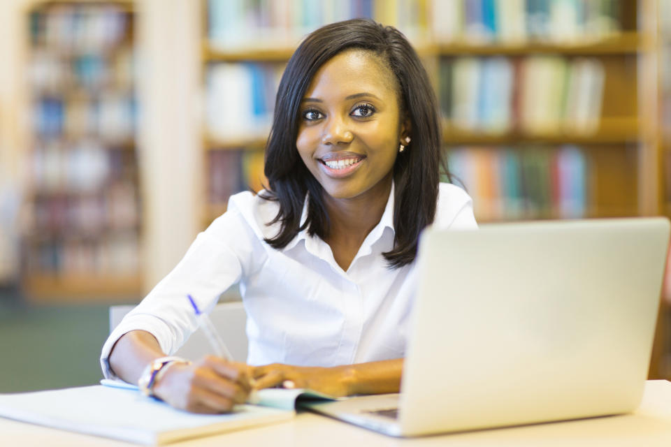 Smiling young woman in library at laptop writing in a notebook