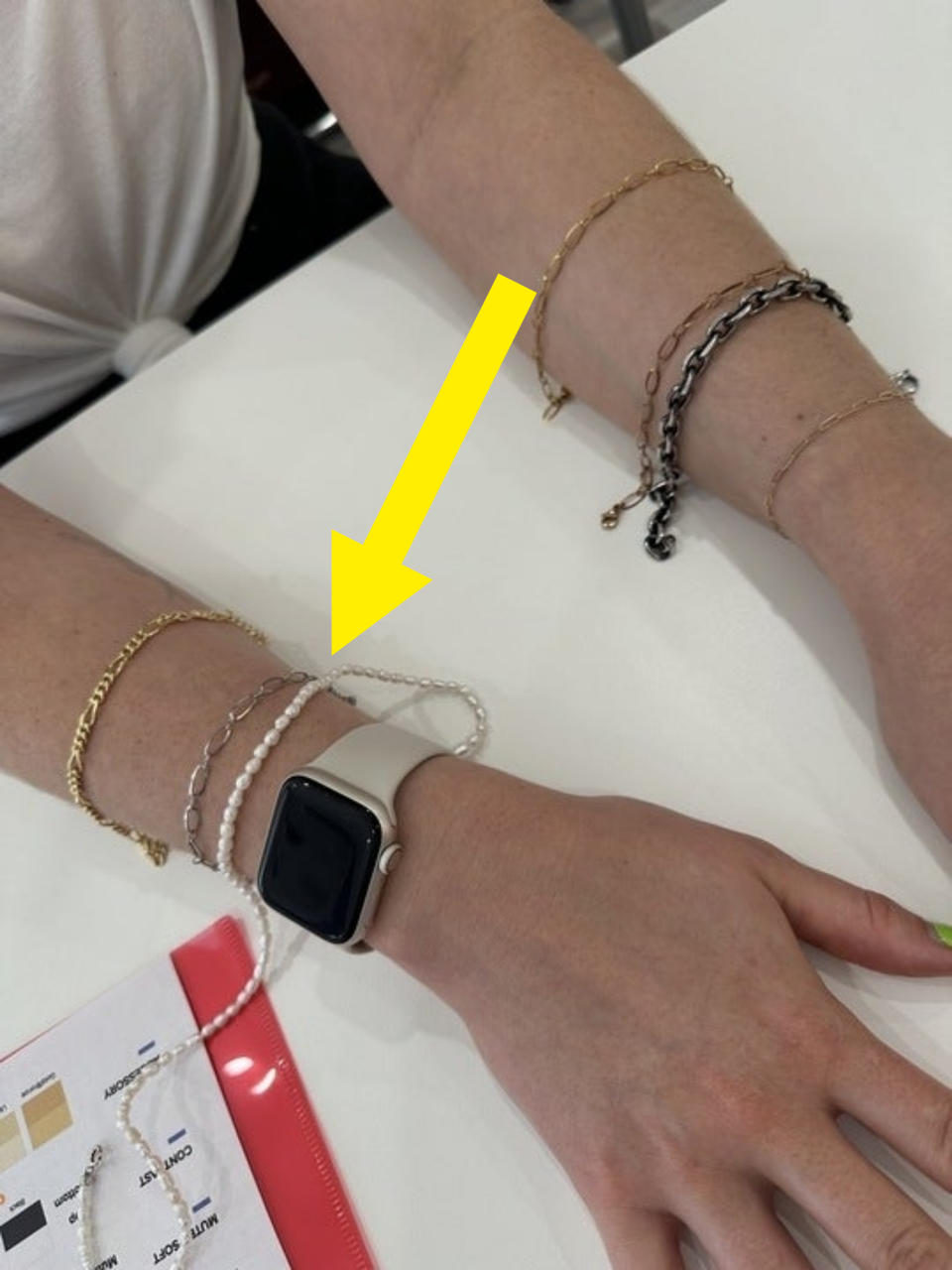 A person wearing multiple bracelets and a smartwatch on their wrists, with a color swatch and paper on the table in front