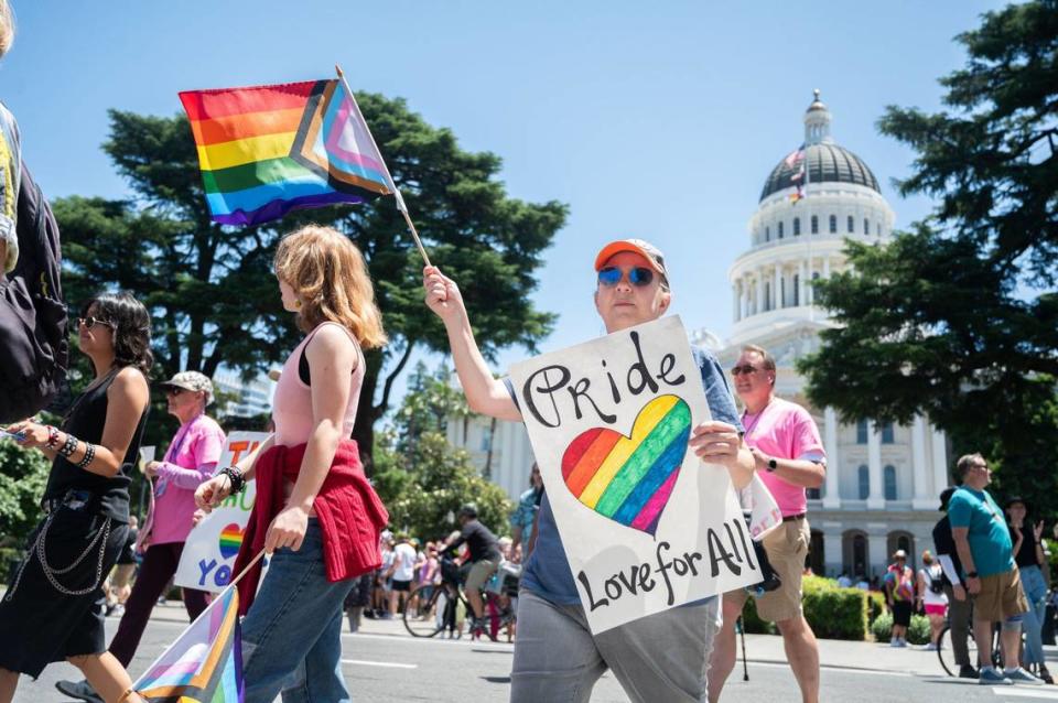 Una participante en la Marcha del Orgullo efectuada  el domingo ondea una bandera de arco iris y sostiene un letrero en el que se lee "Orgullo, amor para todos" mientras camina frente al capitolio del estado en el centro de Sacramento. Algunos asistentes que no participaban en el desfile se ofrecieron a dar abrazos gratis a la gente.