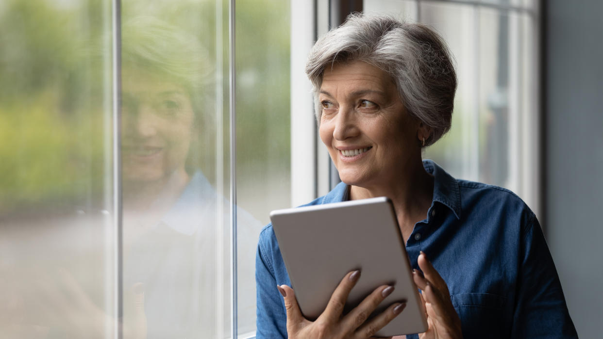 Attractive middle-aged woman holding tablet looking out the window. On-line recreational resources e services for older citizen, e-health remote counselling, e-commerce satisfied mature user concept