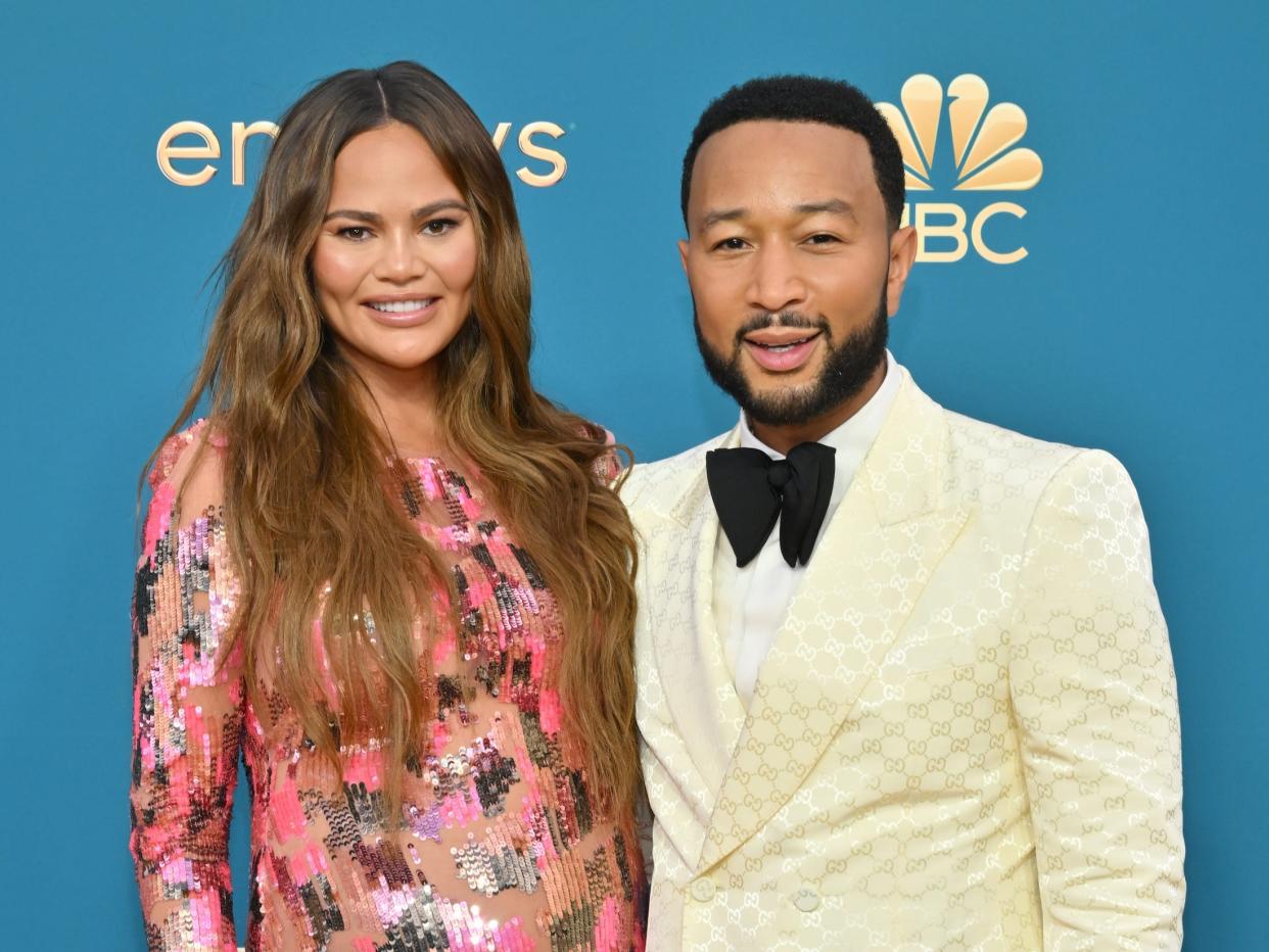 Chrissy Teigen, in a pink sparkly gown, poses with John Legend, in a cream suit and black bow tie, on the red carpet.