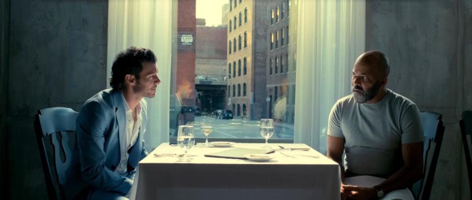 Two men at a restaurant table, one in suit jacket, the other looking tough in a T-shirt.