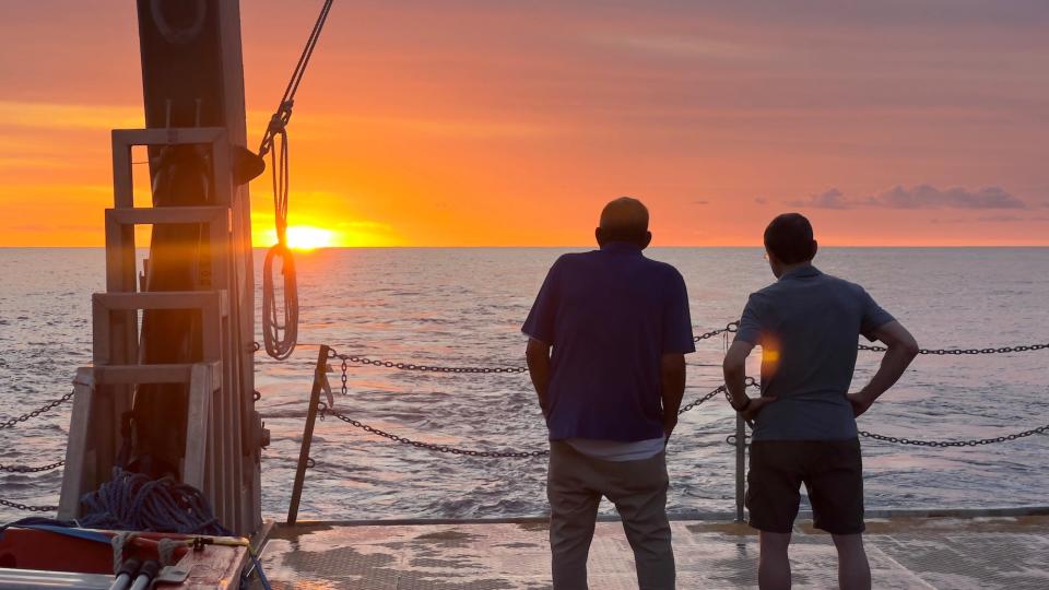 A picture shows Loeb and another person seen from the back on the boat of the interstellar salvage expedition, silhouetted against a sun setting over the sea on the horizon.