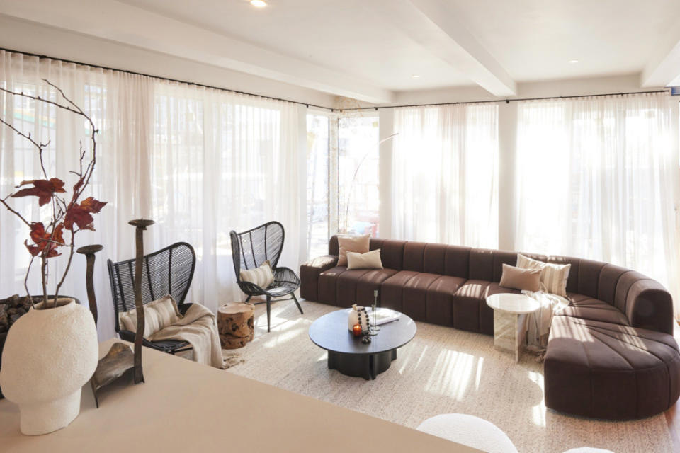 A wide shot of the living room with a large brown lounge, a vase on the left hand side and sheer white curtains all around.