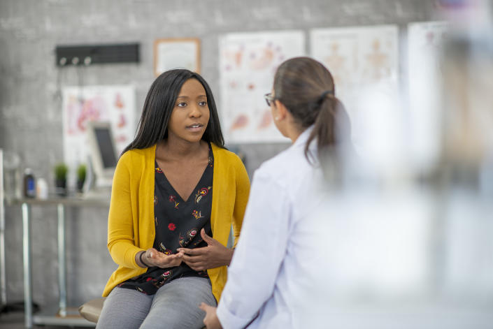 A woman of African descent and her doctor are indoors in a medical clinic. The woman is sitting and describing her symptoms to the doctor.