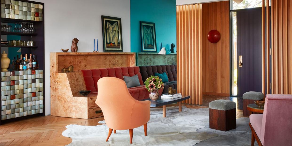 in a midcentury beach house living room is a bar  fronted with colored tiles, a sofa with a burl wood frame and tufted terra cotta cushions, two modern chairs, cocktail table, too stools, and a cow hide rug