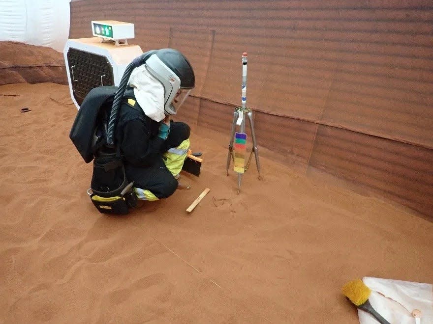 person kneels down in a black spacesuit with a helmet and oxygen backpack looking at a tripod in a room filled with red sand